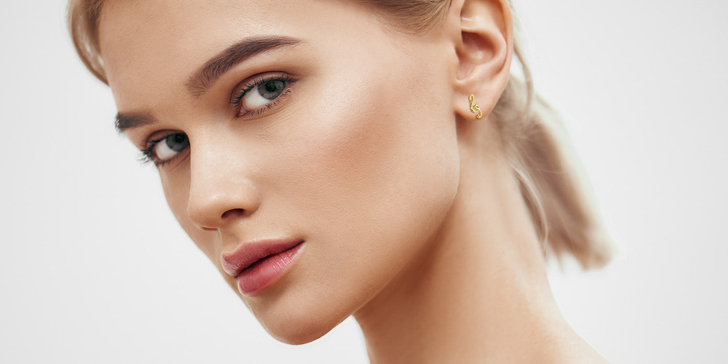 Elegante junge Frau mit blonden Haaren trägt zierliche goldene Ohrringe mit geschwungenem Notenschlüssel, natürliches Make-up mit betonten Augen und rosigen Lippen, Porträt vor weißem Hintergrund