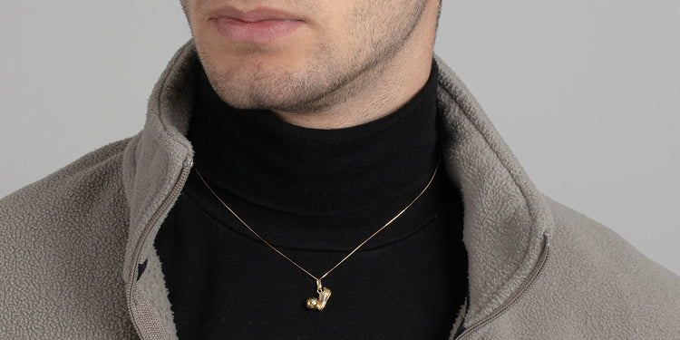 Mann trägt goldene Halskette mit Fußball-Anhänger zu schwarzem Rollkragenpullover und grauer Jacke - eleganter Herrenschmuck