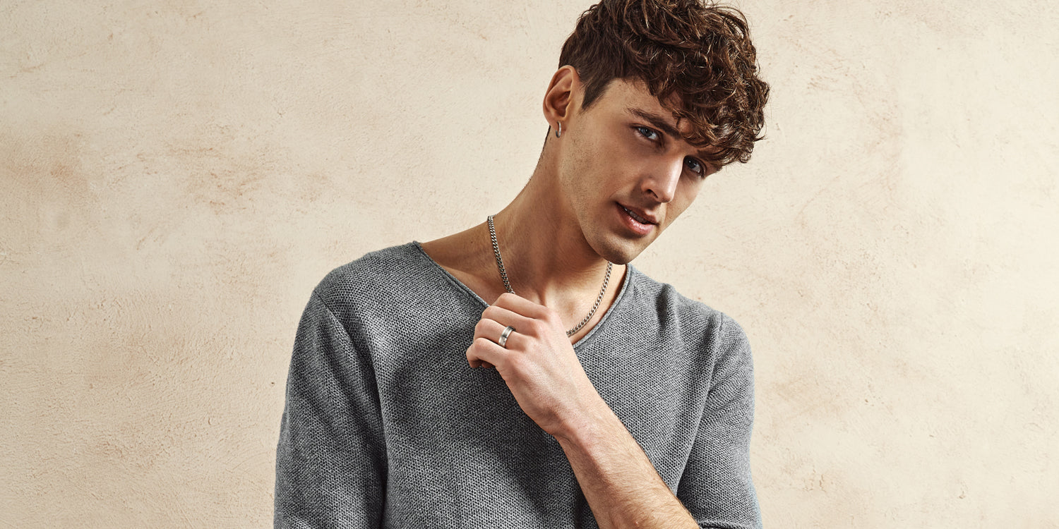 Junger Mann mit lockigen Haaren trägt goldene Panzerkette und silbernen Ring in grauem Pullover