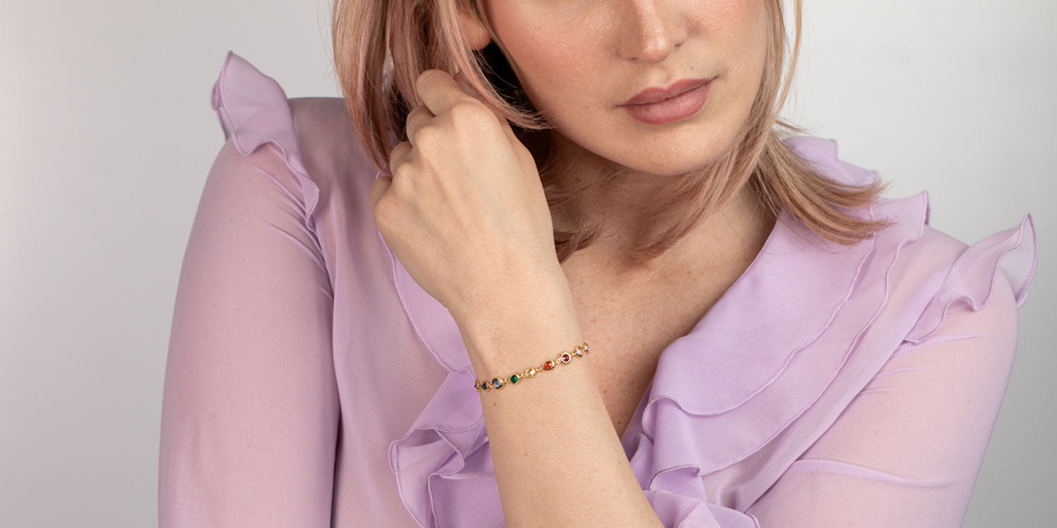 Frau in lila Rüschenbluse trägt goldenes Armband mit bunten Edelsteinen am Handgelenk