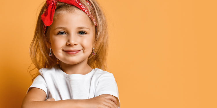 Lächelndes Mädchen mit blonden Haaren und rotem Haarband trägt bunte Ohrringe zu weißem T-Shirt vor orangem Hintergrund - fröhlicher Kinderschmuck