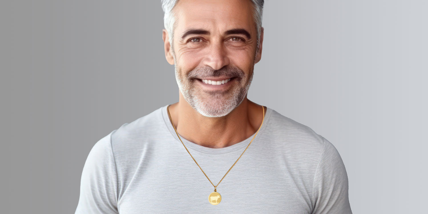 Lächelnder Mann mit grauem Bart in weißem T-Shirt trägt goldene Halskette mit rundem Anhänger mit Kuh-Motiv 