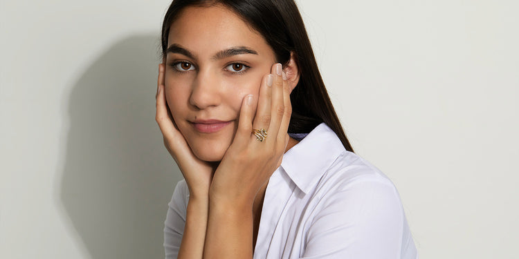 Junge Frau mit dunklen Haaren stützt Gesicht in die Hände und trägt einen auffälligen eleganten Ring am Finger, dazu ein weißes Hemd