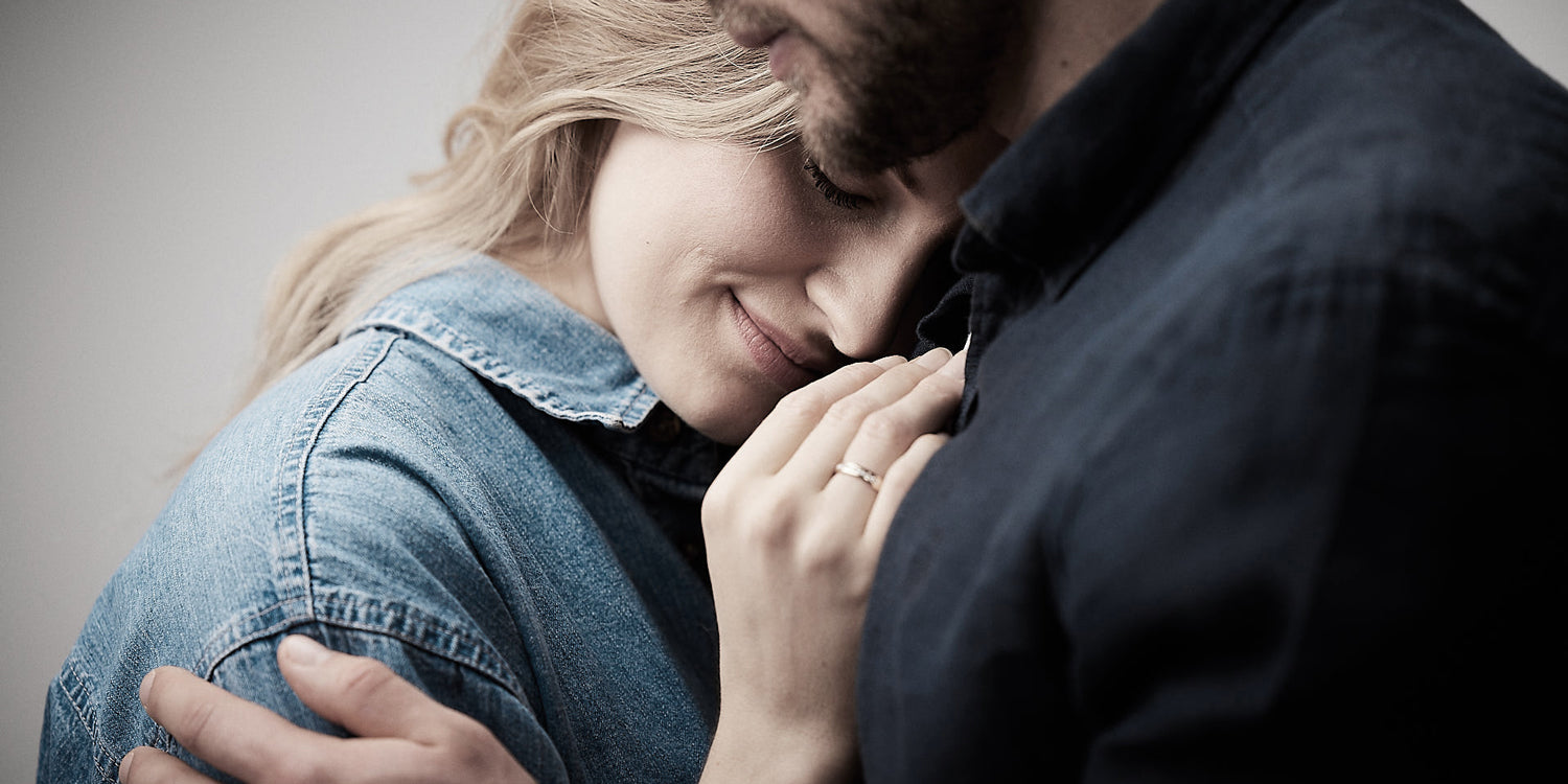 Ein Paar in einer innigen Umarmung, eine blonde Frau in Jeansjacke und mit edlem Fingerring lehnt sich an einen Mann in dunkler Kleidung