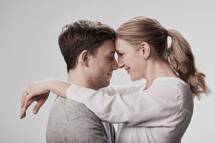 Romantisches Paar in inniger Umarmung - beide tragen passende Ringe, Mann in grauem Pullover, Frau in weißer Bluse